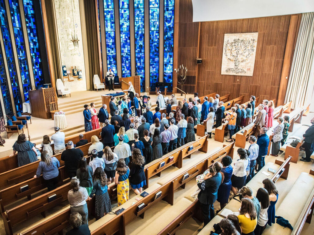Congregation stands in a church sanctuary with blue stained-glass walls and a speaker at the lectern near the altar.